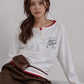 ALYSI ホワイトの長袖ヘンリーネックTシャツ、赤いインナーを重ねた座り姿と胸元のメッセージプリント