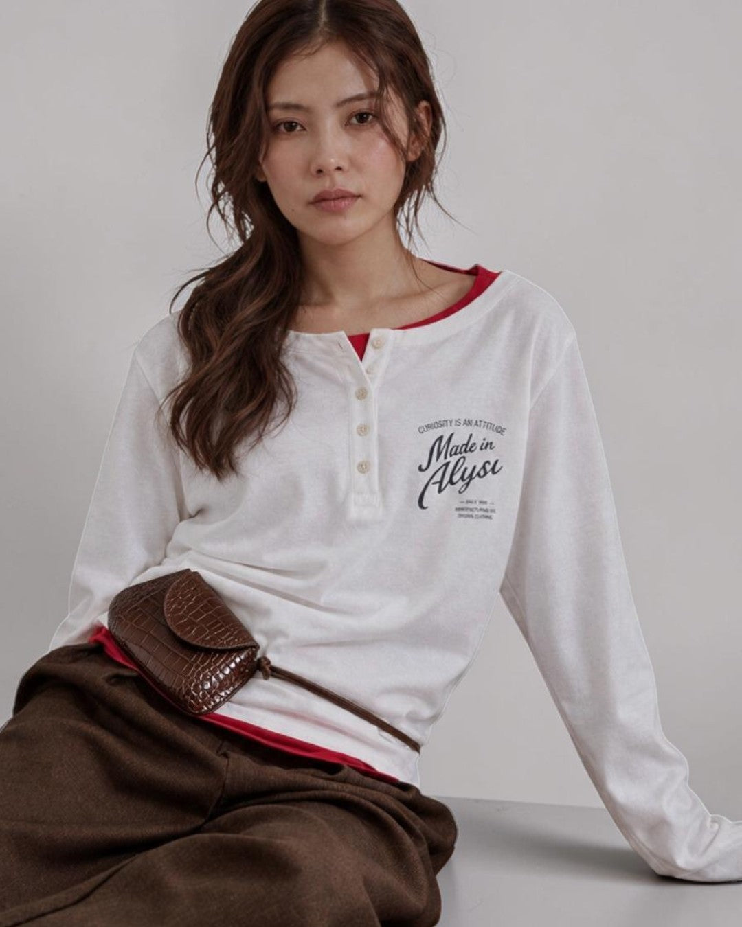 ALYSI ホワイトの長袖ヘンリーネックTシャツ、赤いインナーを重ねた座り姿と胸元のメッセージプリント