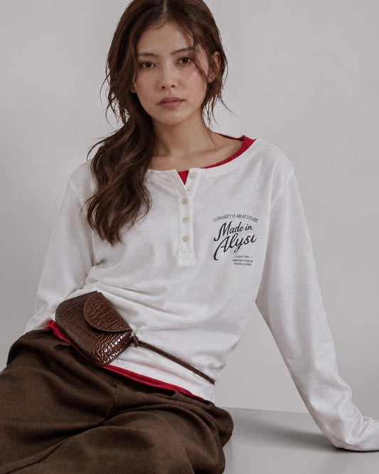 ALYSI ホワイトの長袖ヘンリーネックTシャツ、赤いインナーを重ねた座り姿と胸元のメッセージプリント